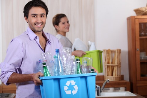 A young man with dark hair and a beard, wearing a light purple shirt, is smiling and holding a blue recycling bin filled with clear glass bottles and jars, in a domestic kitchen. Behind him, a woman with long brown hair, dressed in a gray top, is standing near a sink, with her arms crossed and a neutral expression. The kitchen features light-colored curtains, wooden cabinets, and a countertop with a small green container. There is natural lighting illuminating the scene, highlighting the cleanliness and organization of the kitchen space. The image reflects a setting related to waste disposal and recycling activities in a residential environment, supporting the context of rubbish management services offered by Commercial Waste Removal Victoria.