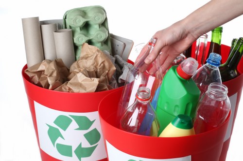 A close-up view of a red recycling bin with a green recycling symbol on the side, filled with various recyclable waste materials. The bin contains crumpled brown paper, several white and brown cardboard tubes, and an empty egg carton made of green plastic. A person's hand is seen reaching into the bin on the right, which holds multiple empty plastic bottles of different shapes and sizes, some with caps, and a few glass bottles with green and red caps. The background is plain white, highlighting the contents of the bins, which are intended for waste collection and recycling services typical of commercial waste removal in Victoria. The scene emphasizes the types of waste commonly handled by professional rubbish removal services, displaying recyclable packaging and bottles in an organized manner.