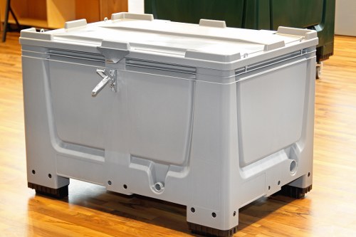 A large, heavy-duty, grey plastic storage container with a secure lid and sturdy construction, placed on a wooden floor indoors. The container has reinforced edges, a locking latch at the front, and visible drainage holes at the base for easy emptying or cleaning. The surface shows a matte finish with some textured patterns to enhance grip and durability. The container appears to be used for storing or transporting bulky or waste materials, aligned with commercial waste management practices. It is positioned against a background featuring cabinets and other storage units typical of a workshop, warehouse, or commercial premises, suggesting its suitability for rubbish or waste removal tasks. Commercial Waste Removal Victoria services are associated with such waste containment solutions in the Victoria area, which includes the local Postcode region. The image focuses on the container’s practical design and functional features intended for efficient rubbish handling in a professional setting.