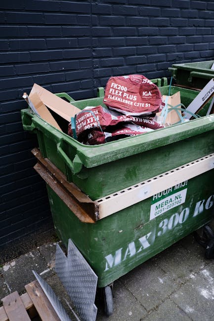 A large green wheeled waste bin filled with bags of building materials, including red bags labeled 'FK200 FLEXY PLUS FLEXLEBER CZTE S1,' situated outdoors on a paved surface against a dark blue brick 
