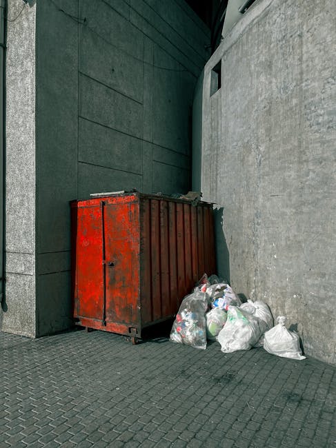 A worker operating a bright red garbage collection truck on a street during daytime, with the rear loading mechanism open and actively collecting waste. The worker, dressed in a yellow and red uniform